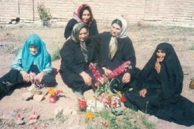 Four women in dark clothing and headscarves sitting around a grave with flowers and a framed photograph.