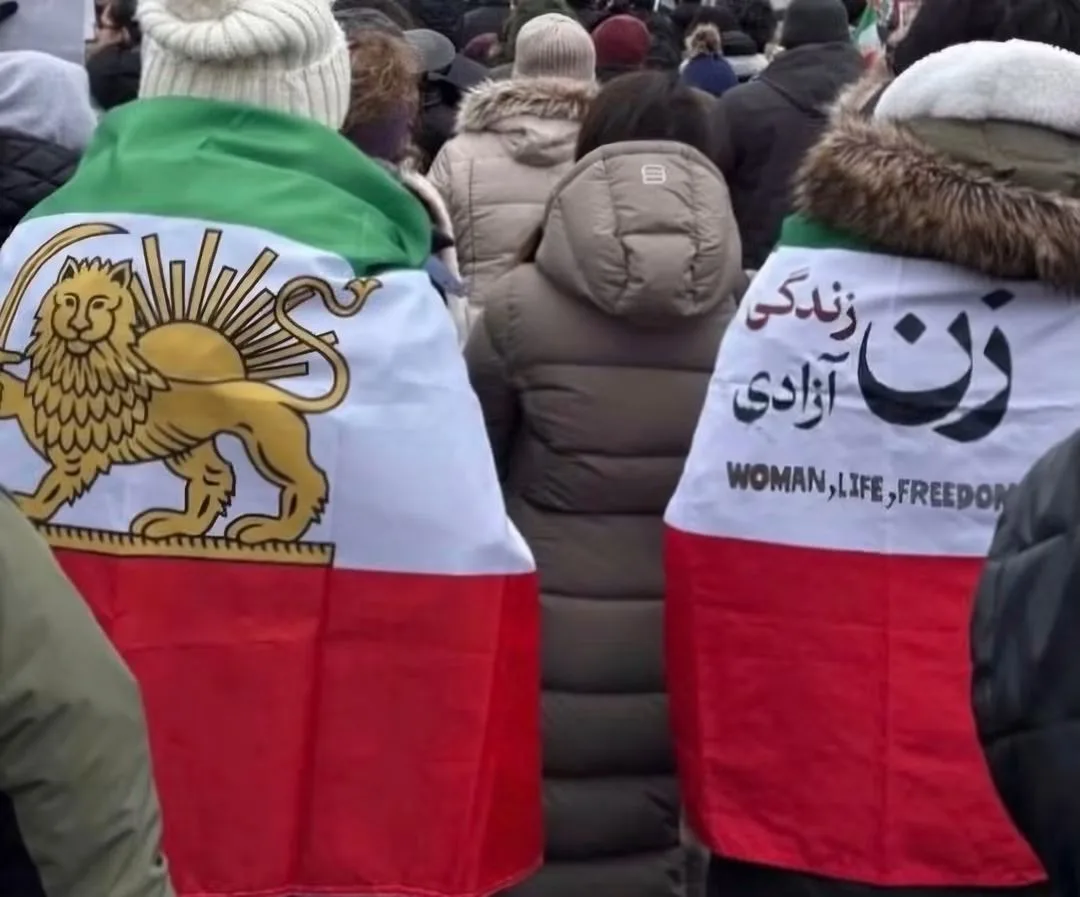 Two people at a winter rally, seen from behind, each wearing an Iranian flag as a cape. One flag carries the Lion and Sun; the other reads Woman, Life, Freedom in Persian and English.